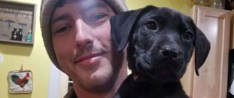 Man in beanie holds black puppy in a kitchen with yellow walls and wooden cabinets, showcasing pet care moment.